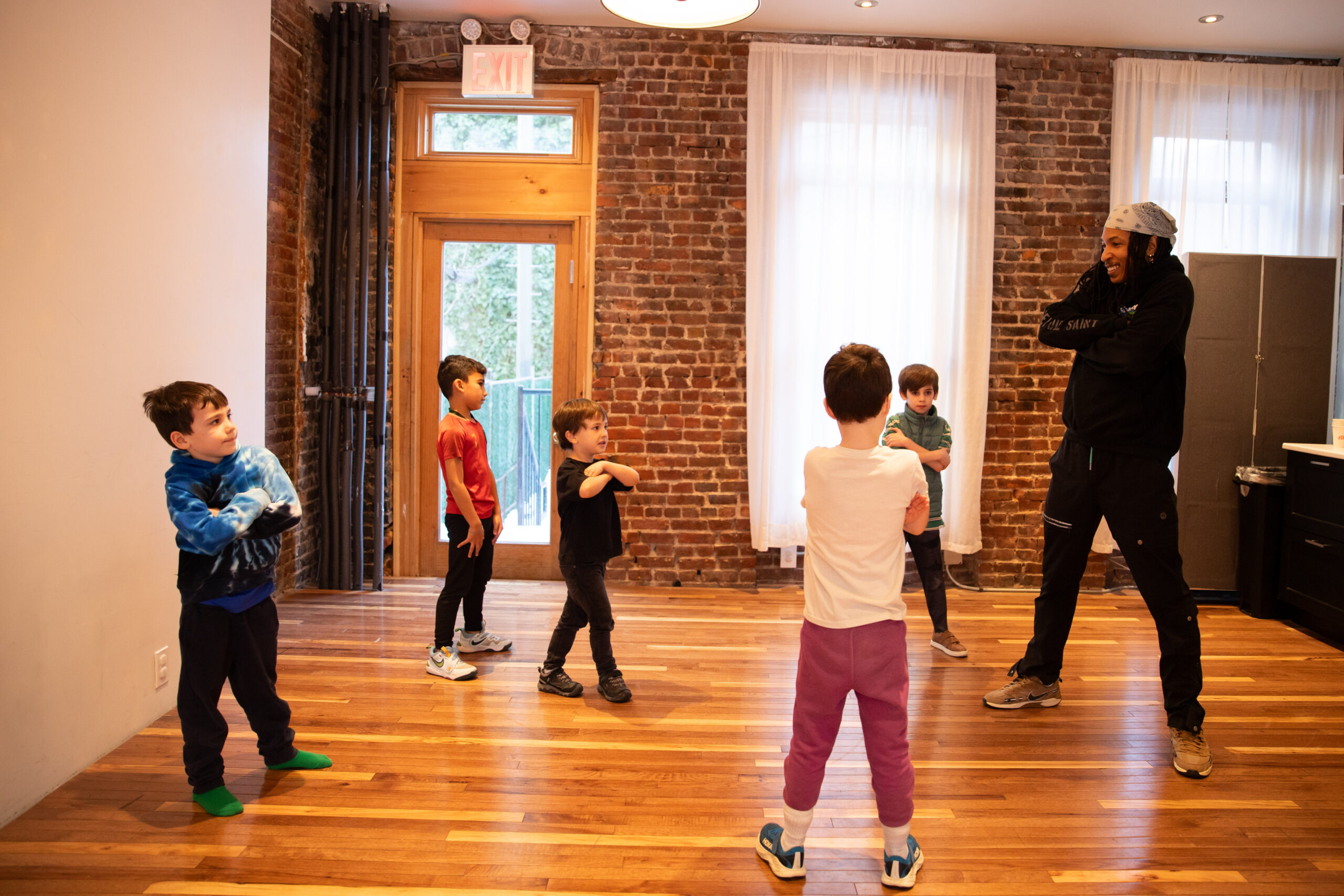 Photo of a dance instructor leading a group of young children practicing a cross-armed movement in a studio with brick walls and wood floors.