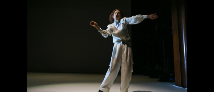 A woman in baggy white clothing dances under a spotlight in a darkly lit room.