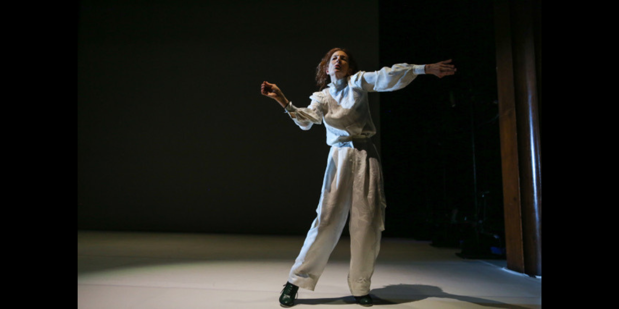 A woman in baggy white clothing dances under a spotlight in a darkly lit room.
