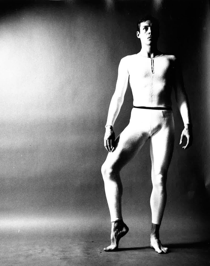 A dancer stands, looking into the distance with one foot in a demi-pointe position. 