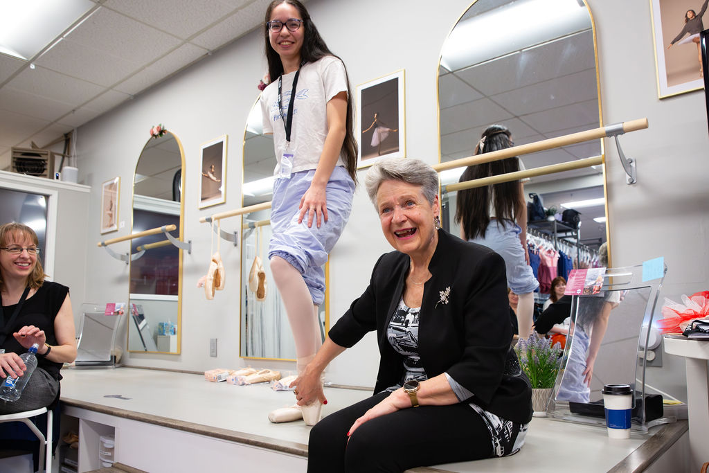 Juon smiles and sits on a raised platform while fitting a dancer for pointe shoes.