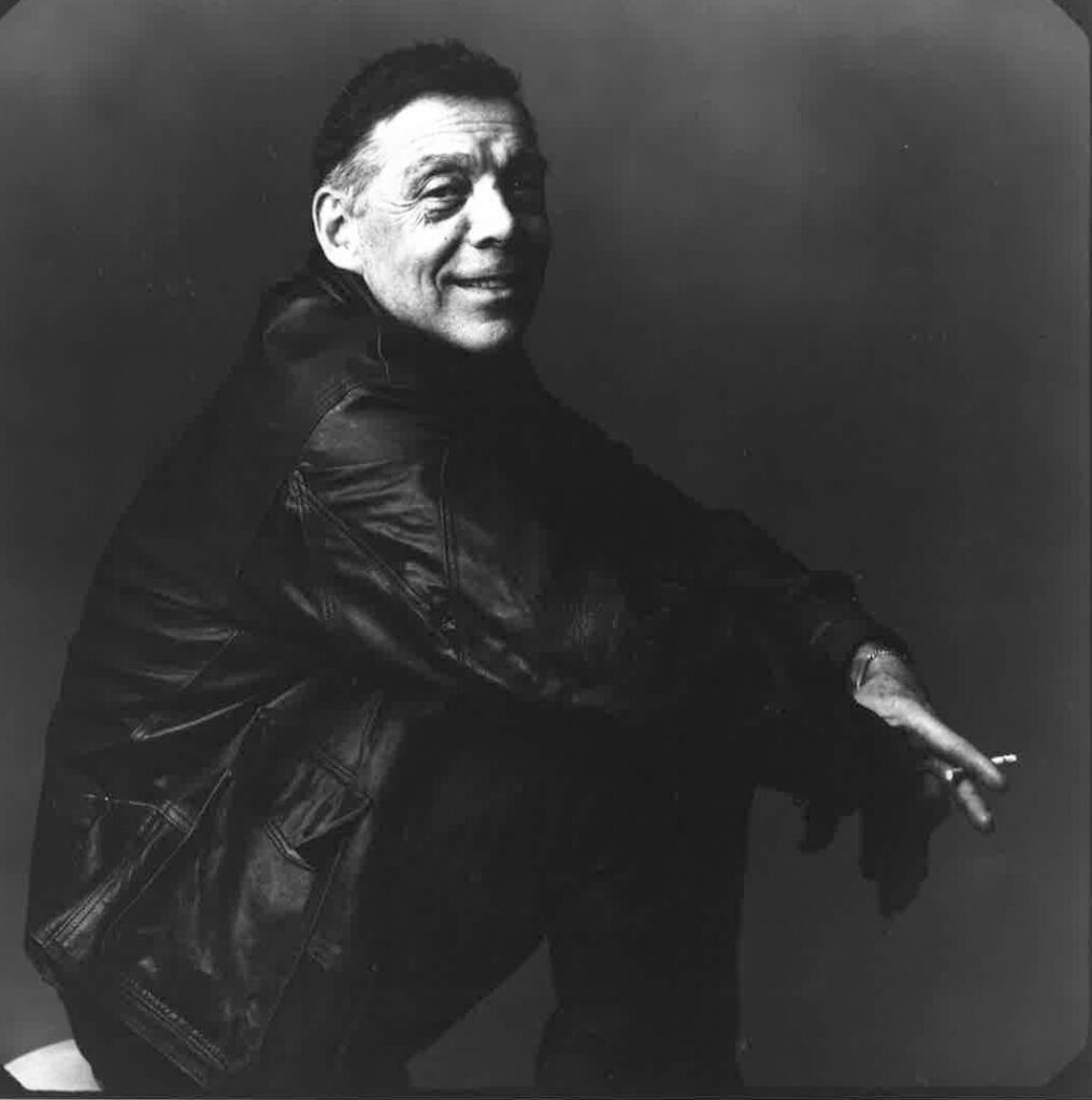 Paul Taylor, wearing black clothing, sits and looks over his shoulder, smiling. 