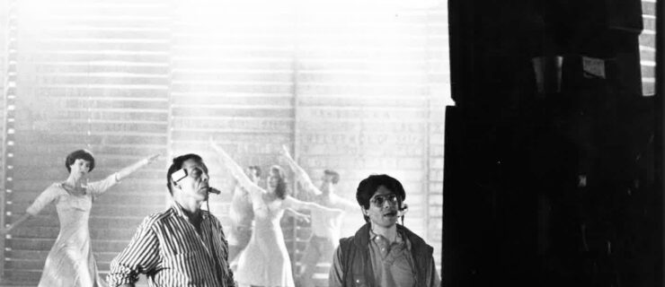 Two men wearing headsets look at a screen while people in light-colored clothing dance behind them.