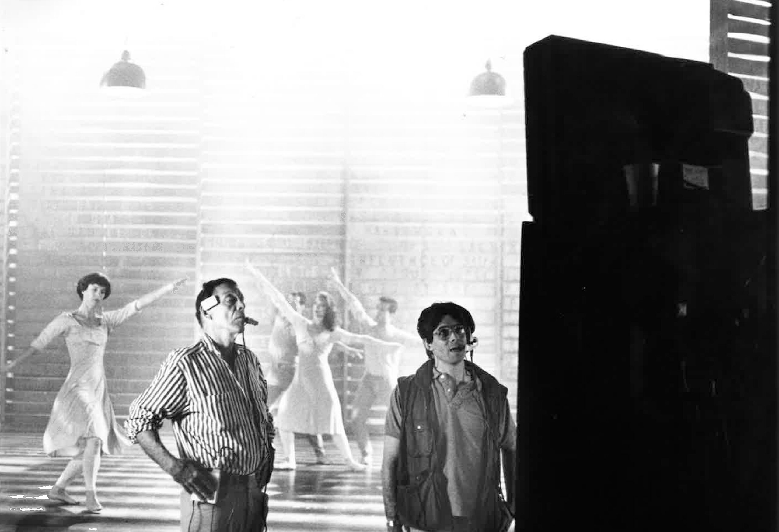Two men wearing headsets look at a screen while people in light-colored clothing dance behind them.