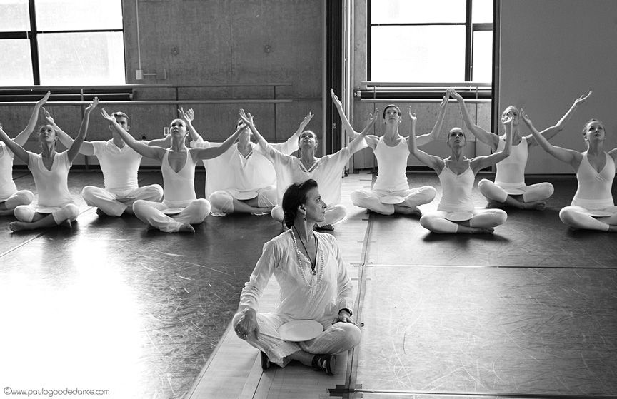 A woman in a loose-fitting white shirt and pants sits crossed-legged on a dance studio floor in front of a staggered line of dancers weering white practice clothes. She looks over her left shoulder at them as they lift their arms high, looking up, and sit crossed-legged on the floor.