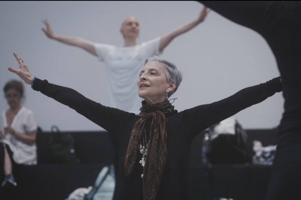A woman with short white hair stands with her arms extended wide, looking out. She wears a long-sleeved black top and a brown scarf tied at her neck.