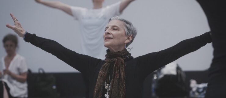 A woman with short white hair stands with her arms extended wide, looking out. She wears a long-sleeved black top and a brown scarf tied at her neck.