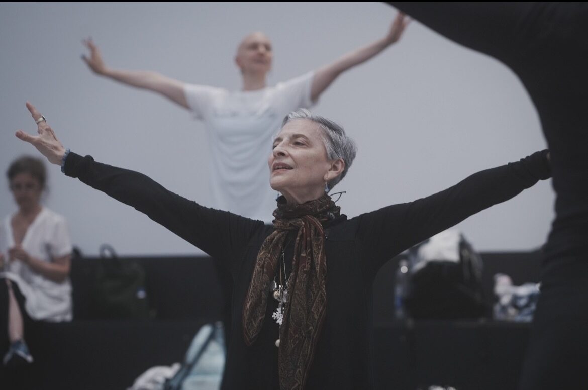 A woman with short white hair stands with her arms extended wide, looking out. She wears a long-sleeved black top and a brown scarf tied at her neck.