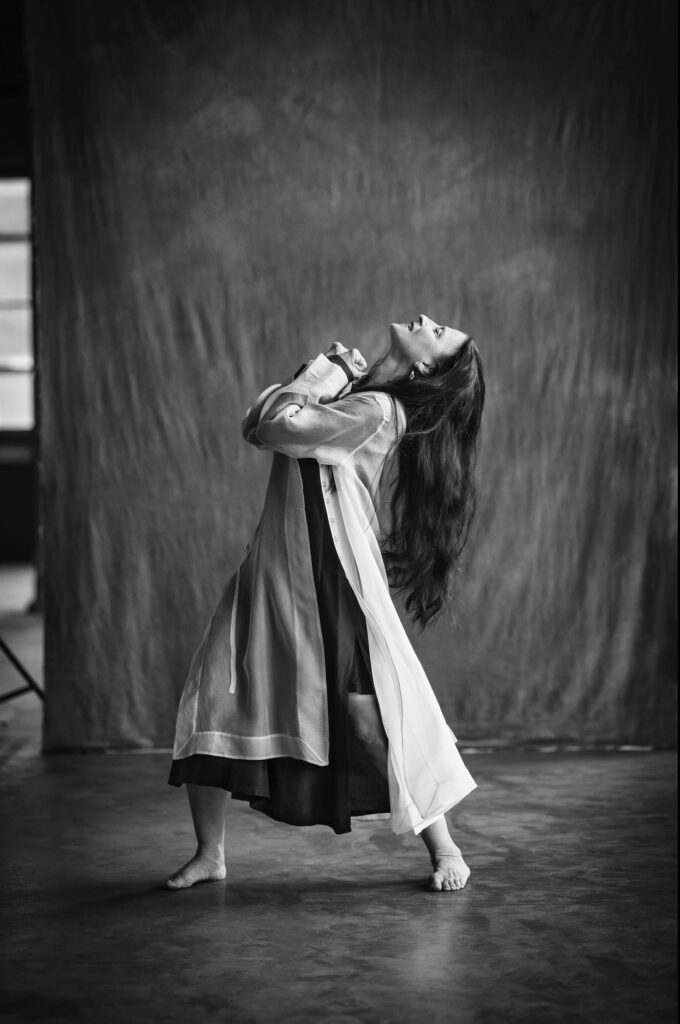 A black and white, full body portrait shows a woman with long dark hair and a loose-fitting, light-colored long-sleeved dress in profile. She stands with her feet in a wide fourth position and clasps her hands to her chest as she looks up, arching back.