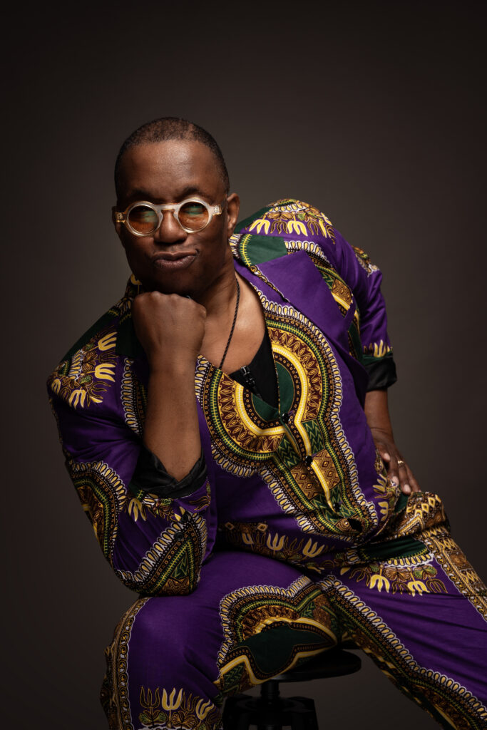 Dancer and teacher Archie Burnett sits facing the camera and rests his chin on his right first. His right elbow rests in turn on his right thigh. He wears transparent-framed sunglasses with circular, yellow-tinted lenses and a purses his lips in a playful manner. His shirt and pants are made of matching fabric printed with multicolored, symmetrical designs on a purple background.