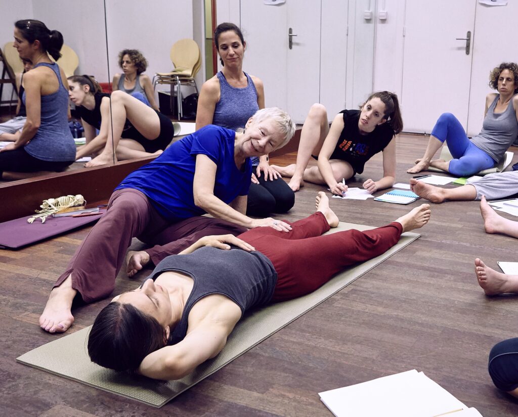 Lynn Simonson assisting a student who is lying down on a yoga mat while other students overlook.