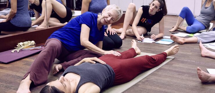 Lynn Simonson assisting a student who is lying down on a yoga mat while other students overlook.