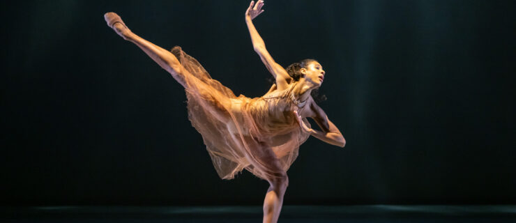 A woman in an orange mesh dress stands in an outreaching arabesque pose on a dark stage.