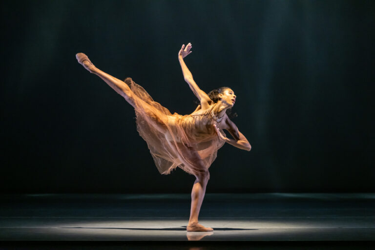 A woman in an orange mesh dress stands in an outreaching arabesque pose on a dark stage.