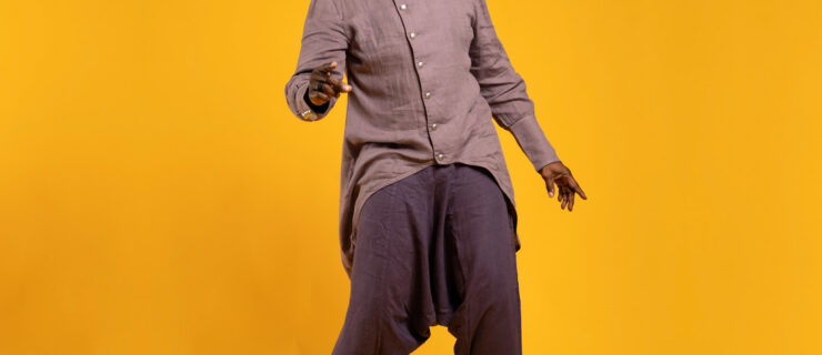 Dancer and teacher Archie Burnett poses with a wide, bright smile in front of a bright-yellow, seamless backdrop. His linen pants are deep purple and his linen shirt is a lighter shade of the same color, closer to lavender. He appears to be caught candidly while dancing.