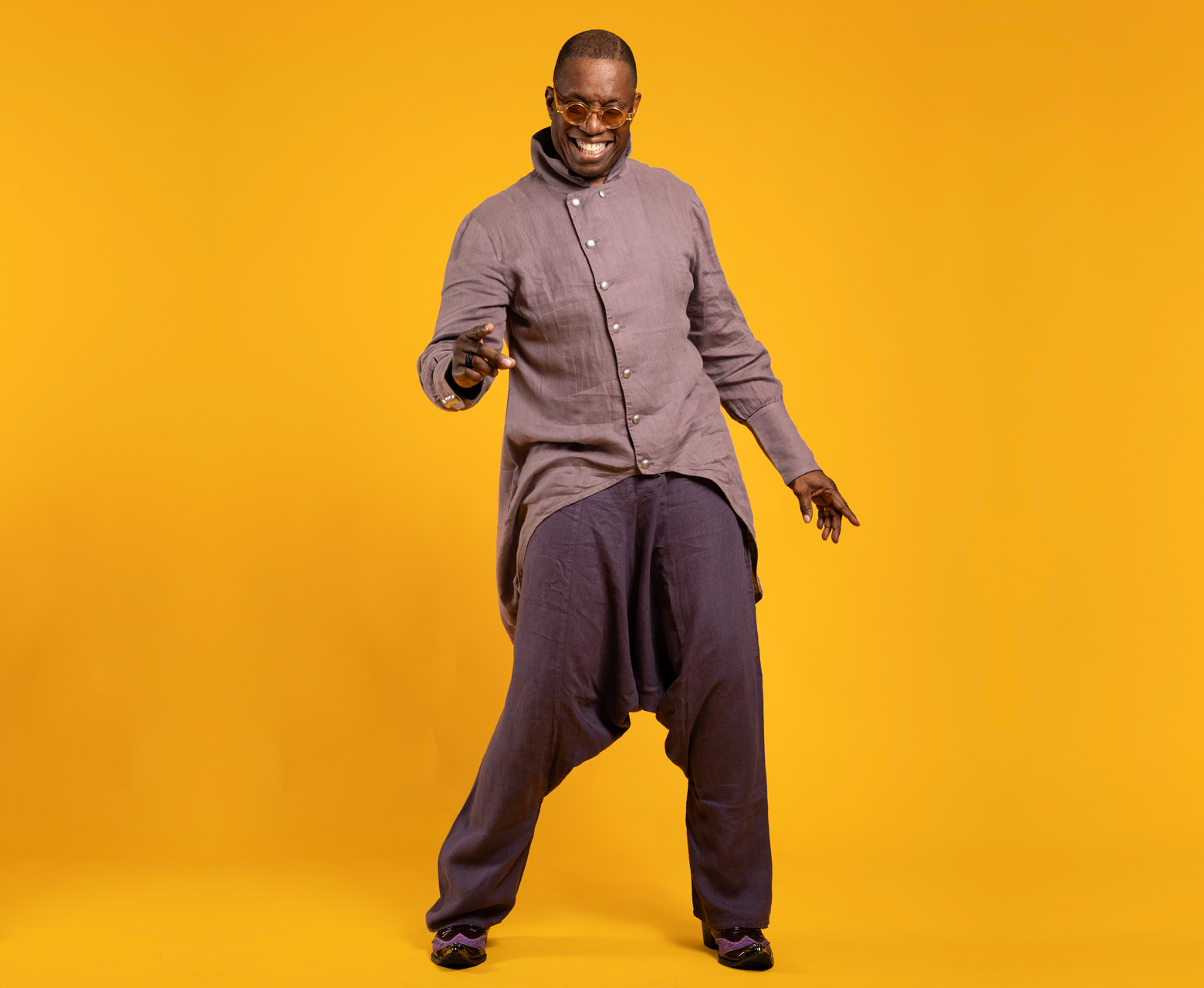 Dancer and teacher Archie Burnett poses with a wide, bright smile in front of a bright-yellow, seamless backdrop. His linen pants are deep purple and his linen shirt is a lighter shade of the same color, closer to lavender. He appears to be caught candidly while dancing.
