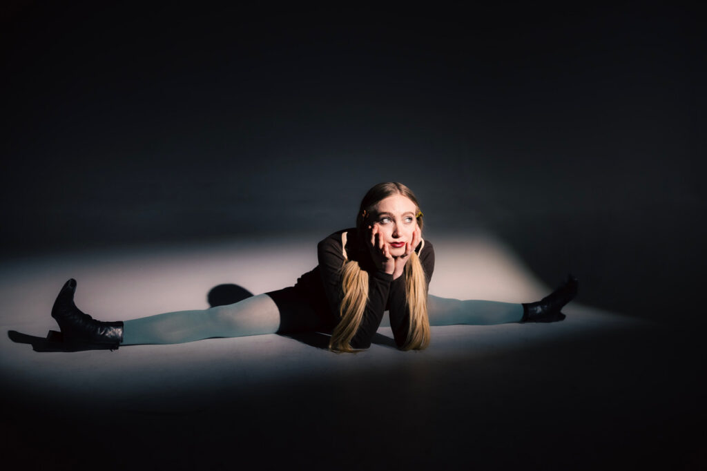 Lucy Vallely in a seated split wearing grey stockings with her face propped on her hands.