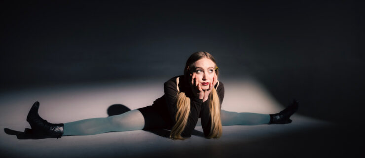 Lucy Vallely in a seated split wearing grey stockings with her face propped on her hands.