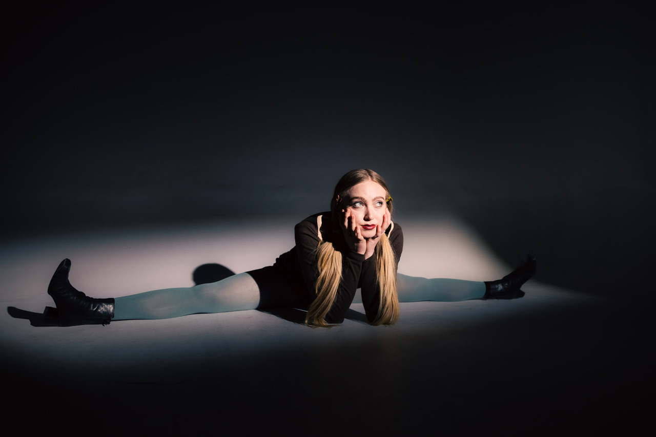 Lucy Vallely in a seated split wearing grey stockings with her face propped on her hands.