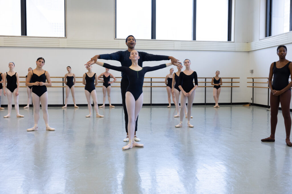In the School of American Ballet studios, Silas Farley holds a female dancers arms out at shoulder height. Farley wears all black. The students wear black leotards and tights.