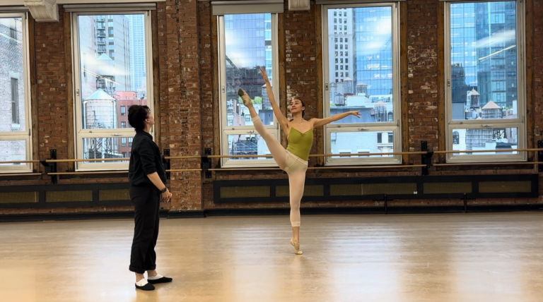 Ballet dancer performing on pointe in the center while a teacher observes in a studio.