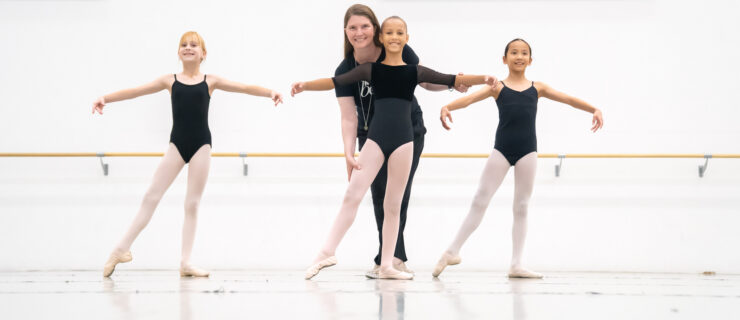 Three young dancers wear black leotards and pink tights as they extend their legs in a tendu to the side in a New Ballet studio with white walls. Dalia Rawson wears all black as she corrects the leg placement of the center girl.