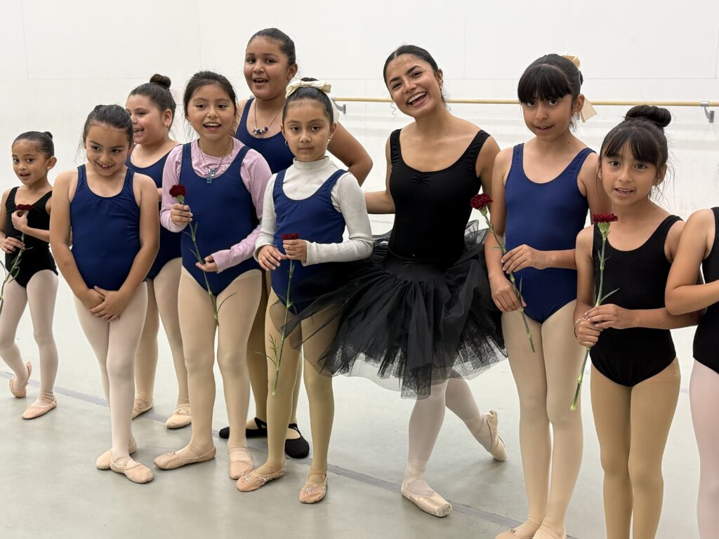 First Step Alum and New Ballet School Continuing Scholarship Student and Intern Dania Barrera wears a black leotard, tutu, and pointe shoes. She has her arms around the 2026 First Step Graduates who all wear navy leotards and ballet shoes in a white New Ballet studio.