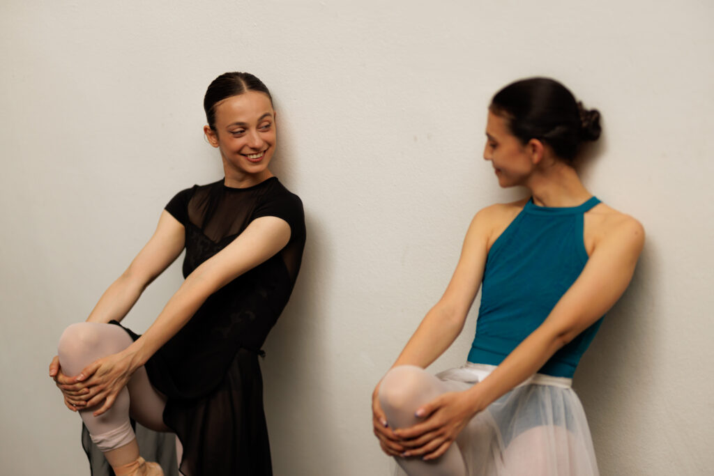 Two ballet dancers stretch and smile together against a studio wall.