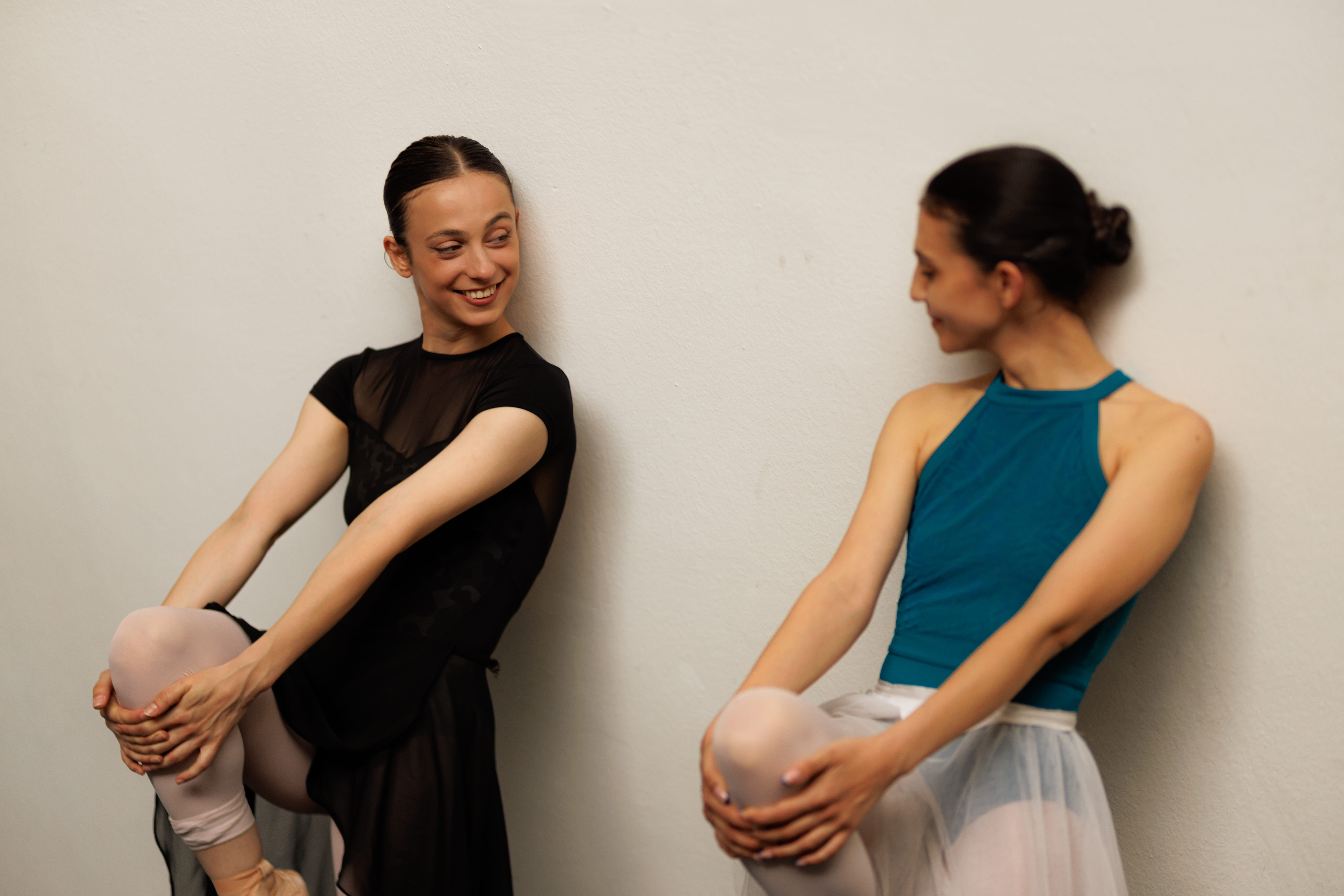 Two ballet dancers stretch and smile together against a studio wall.