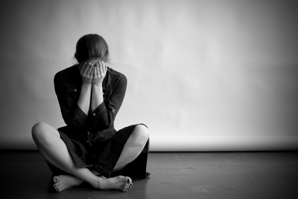 Depressed woman in monochrome sits with her face in the hands and crossed-legged on the floor.