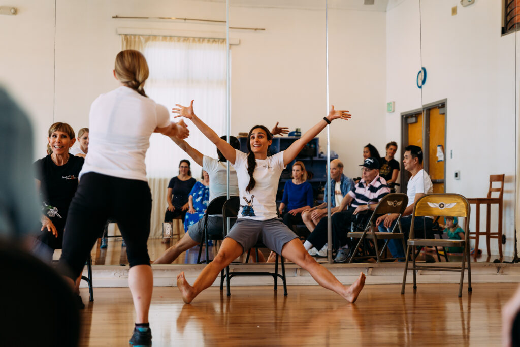 Haylee Nichele teaches a Dancing Through Parkinson's class for Invertigo Dance Theatre. She sits in a chair, smiling, with arms and legs spread wide. In the mirror behind her, you can see class participants seated, watching her. A woman standing points at Nichele with both arms.
