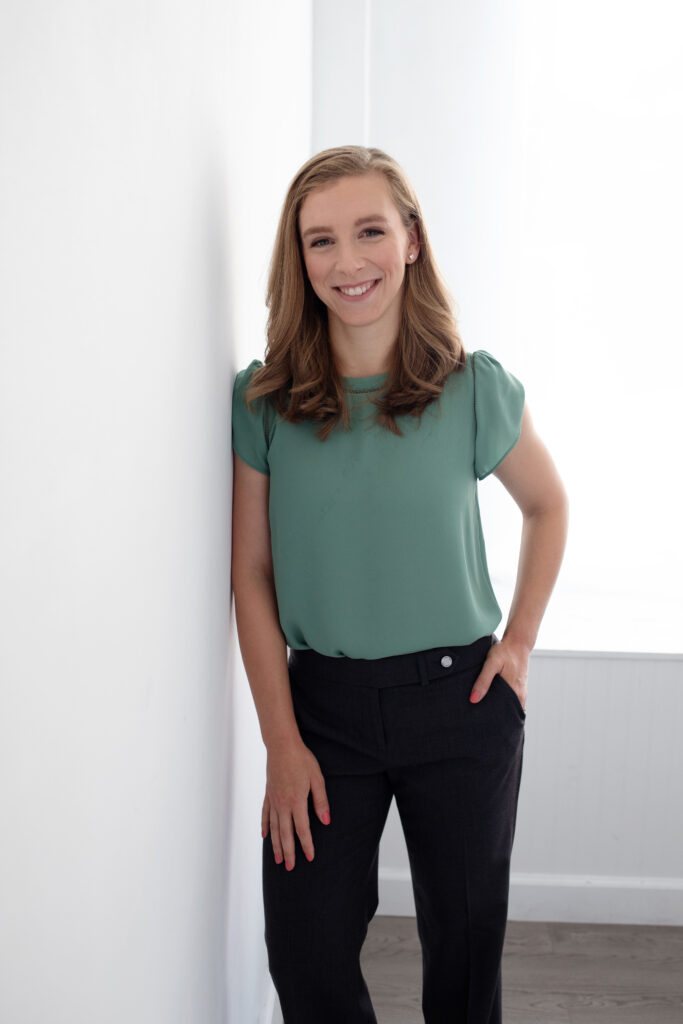 Leigh Skvarla leans against a wall with one hand in the pocket of her black pants. She is smiling and wearing a blue blouse.