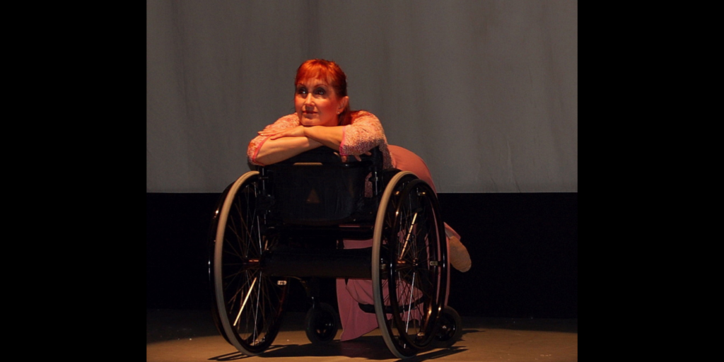 Kitty Lunn seated on a wheelchair in reverse with her face propped over her crossed arms looking outward.