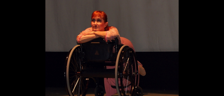 Kitty Lunn seated on a wheelchair in reverse with her face propped over her crossed arms looking outward.
