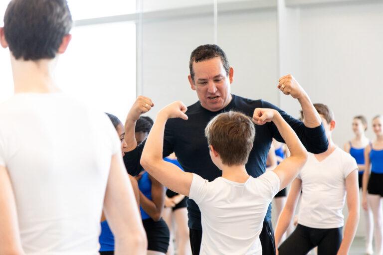 Claudio Muñoz faces a young male dance student whose back is to the camera. They both raise their arms to show their muscles.
