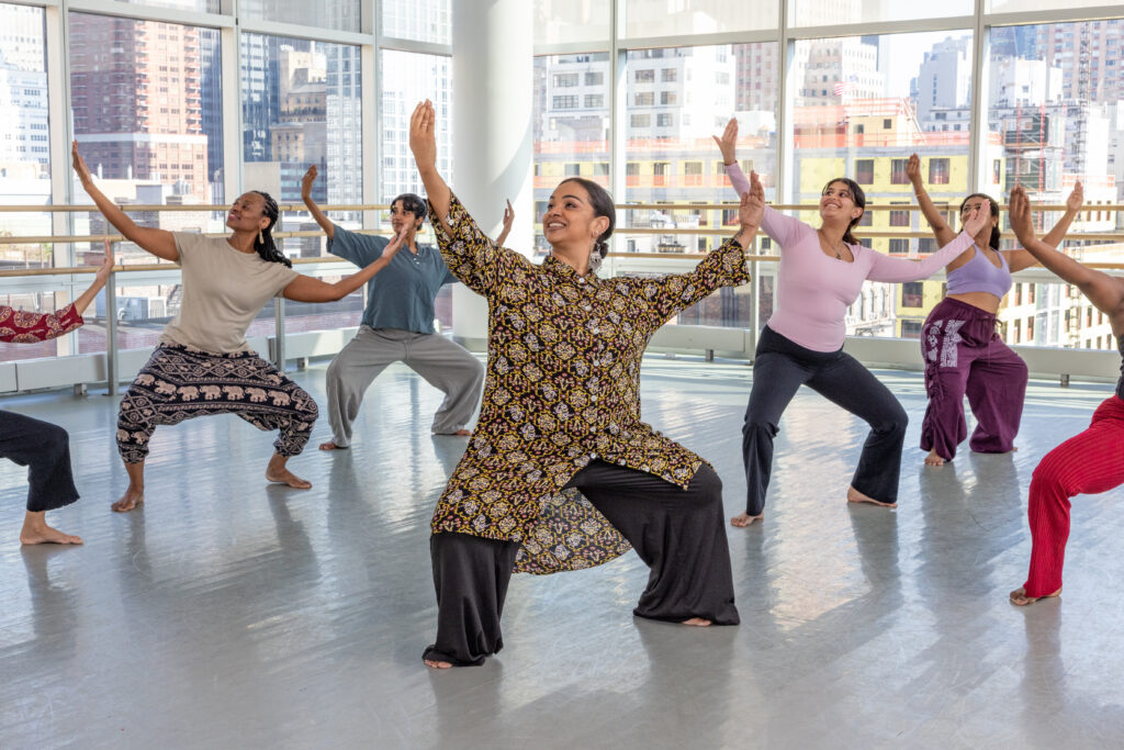 Brinda Guha, wearing brown and black, holds a wide position with legs bent in front of other students holding the same position in a dance studio.