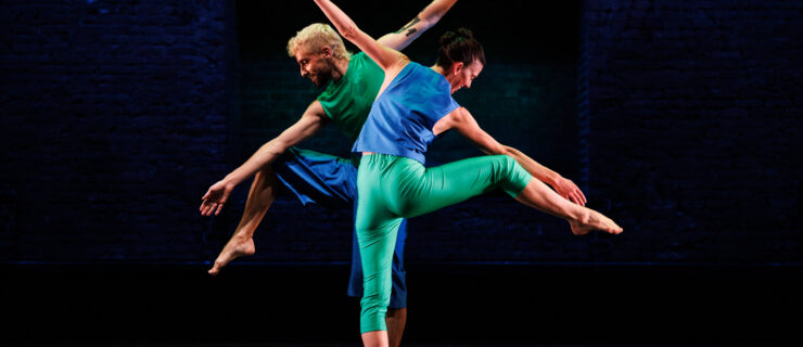 Cecily Campbell and a fellow dance wear blue and green pants and shirts while standing on one leg with arms extended, reaching long. They stand against a dark backdrop.
