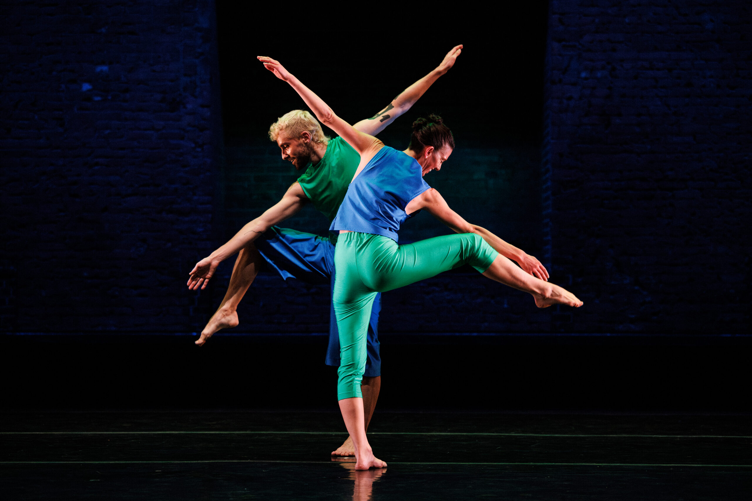 Cecily Campbell and a fellow dance wear blue and green pants and shirts while standing on one leg with arms extended, reaching long. They stand against a dark backdrop.