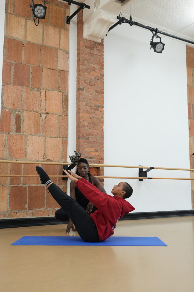 A woman in exercise clothing performs a V-up with her legs and arms extended in front of her. She sits on a blue yoga mat in a dance studio. Her pilates instructor kneels behind her.