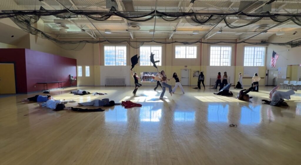 A large group of K-12 students dance in a school gymnasium. Some lay on the ground while others swing jackets in the air.