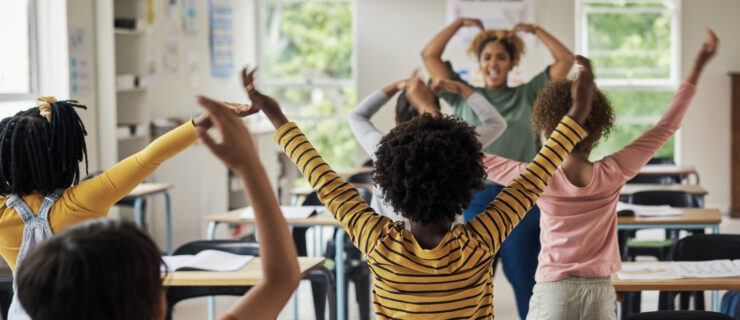 A school teacher leads young students in a dance exercise with their arms above their head in a brightly lit classroom.