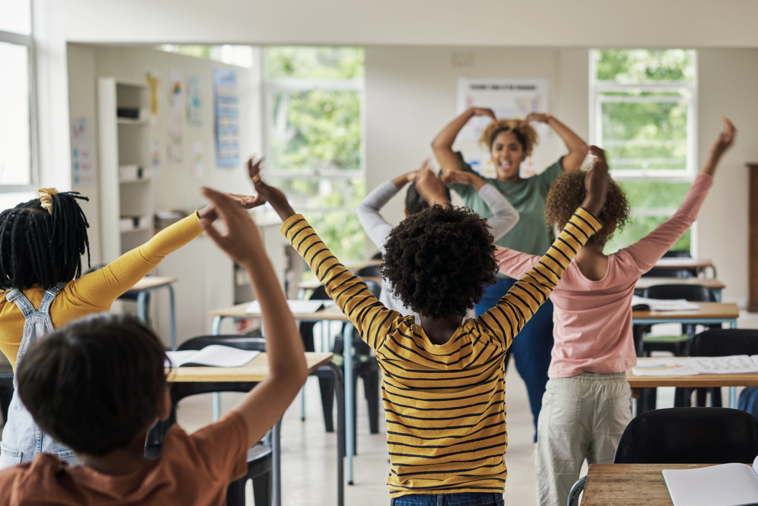 A school teacher leads young students in a dance exercise with their arms above their head in a brightly lit classroom.