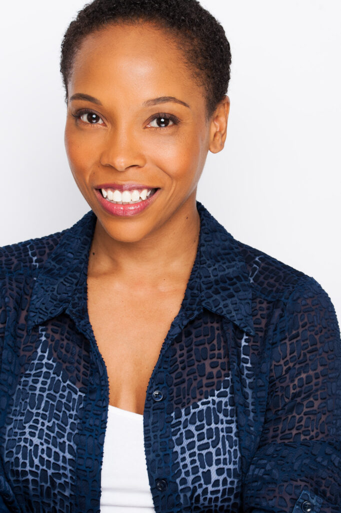 Dionne Figgins smiles for a headshot wearing a blue blouse over a white camisole against a white backdrop.