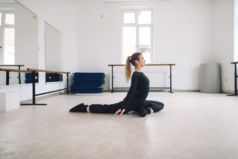 Woman dressed in black dancewear seated in a yoga pigeon pose in a bright, minimalistic dance studio with mirrored walls and ballet barres.