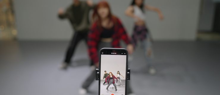 Students rehearsing in a studio (at what looks like a hip hop class) while being filmed by a phone camera set up on a tripod.