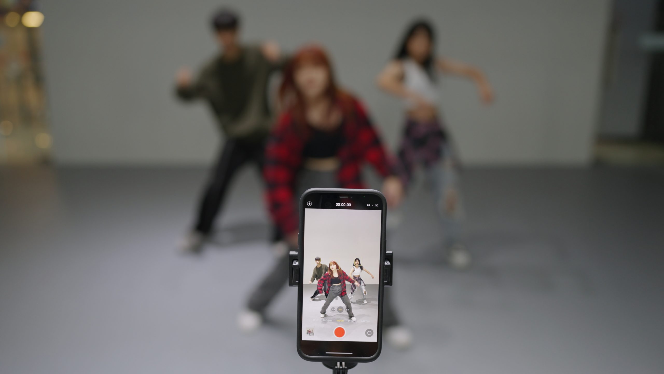 Students rehearsing in a studio (at what looks like a hip hop class) while being filmed by a phone camera set up on a tripod.