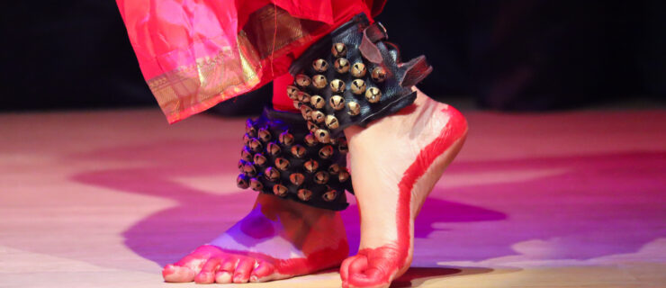 Only the feet of a bharatanatyam dancer are visible. The feet are crossed and the dancer wears pink pants, black ankle cuffs, and red paint on their bare feet.
