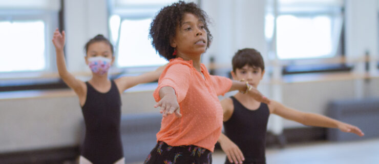 Dionne Figgins wears a coral blouse and demonstrates movement for two young students who are watching her in the dance studio, wearing black ballet uniforms.