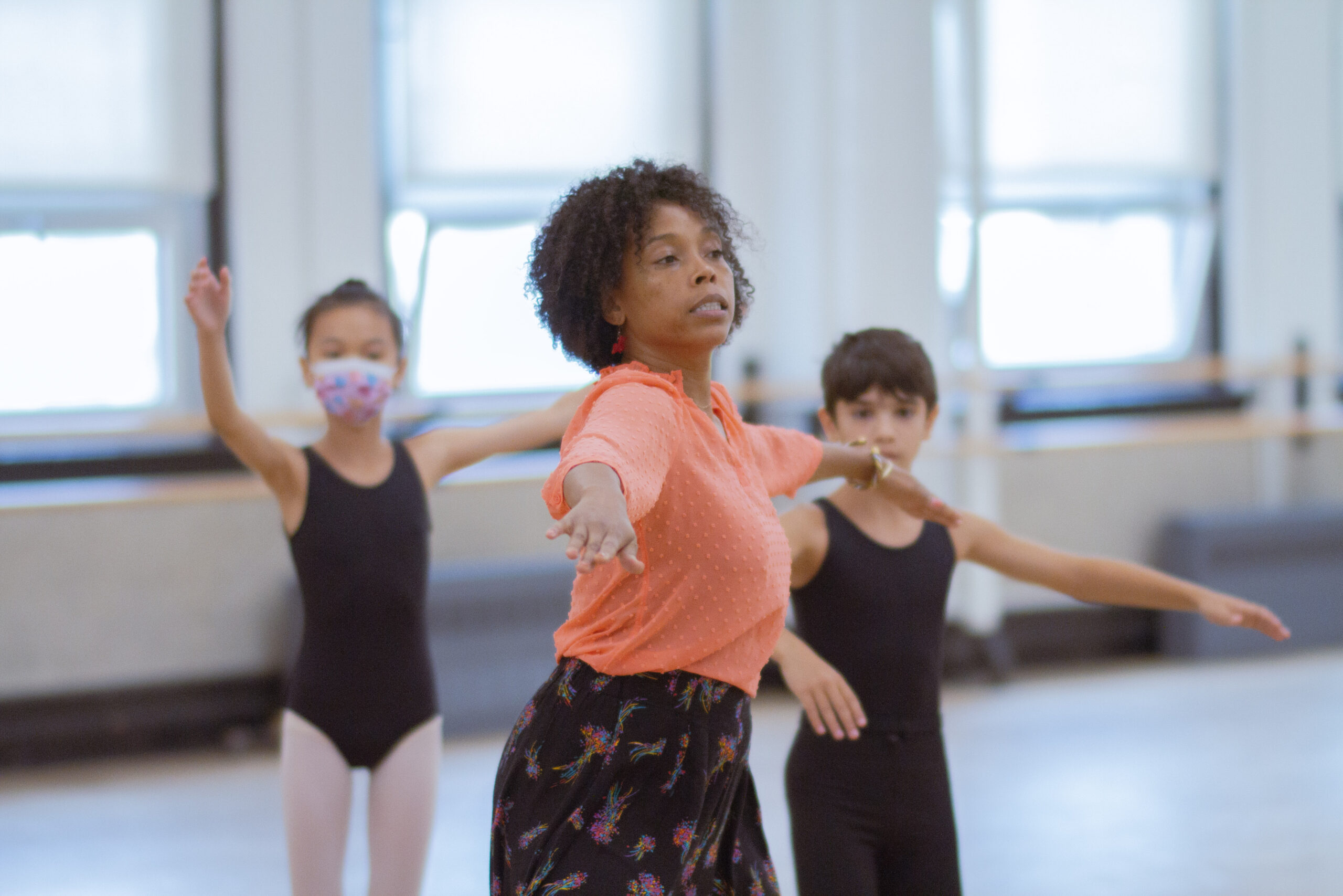 Dionne Figgins wears a coral blouse and demonstrates movement for two young students who are watching her in the dance studio, wearing black ballet uniforms.
