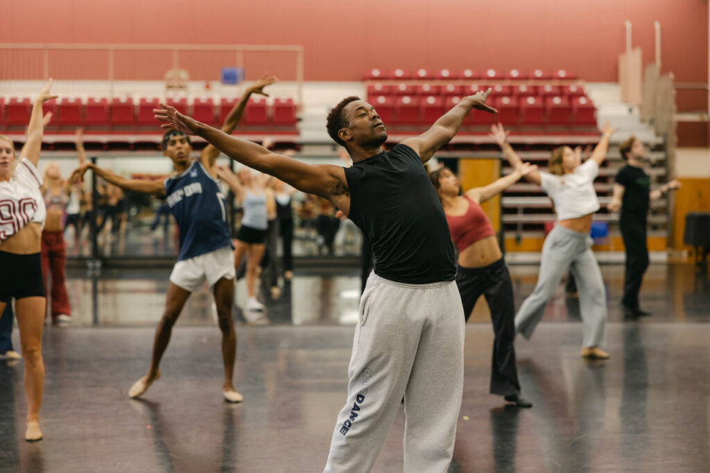 Marquez dances in front of students in a studio. Wearing sweatpants and a tank top, he leans back with his arms extended above his head.